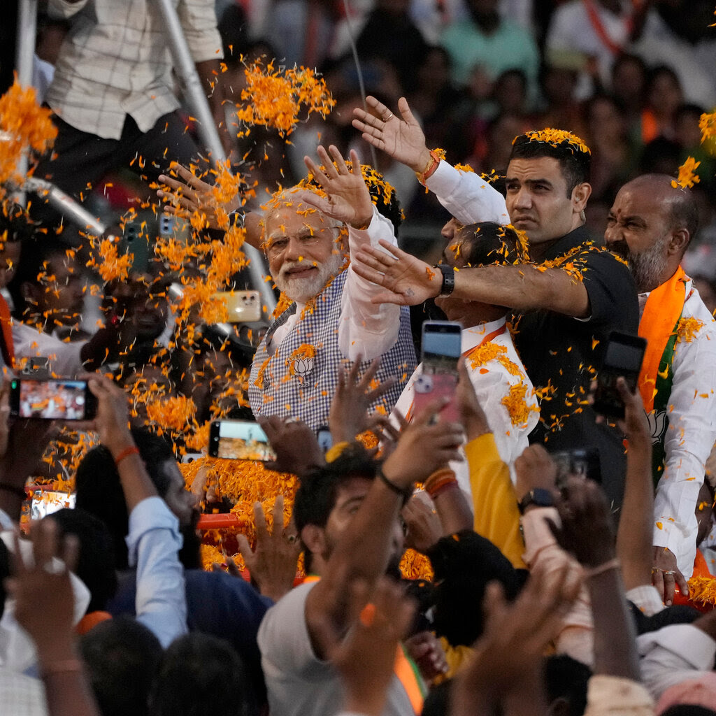 Orange streamers and confetti float over a crowd of people surrounding Narendra Modi waving at a campaign rally.