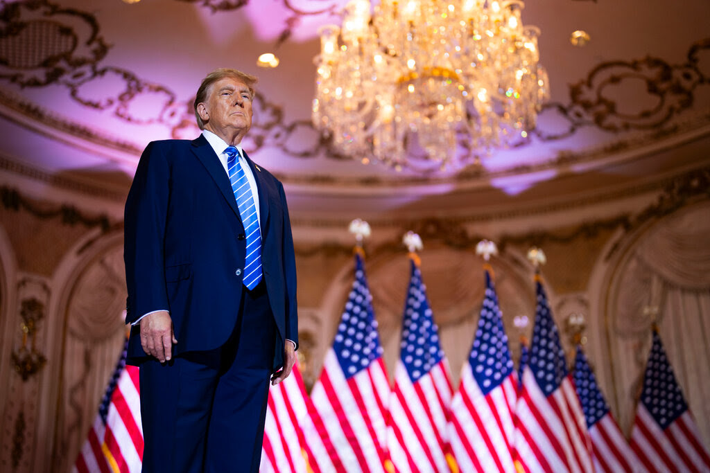 Former President Donald Trump stands in front of a row of flags with a large chandelier above him.
