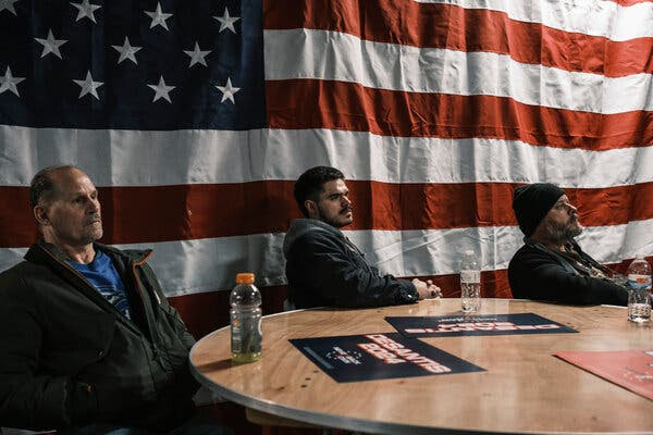 Three men sit at a round table in front of an America flag. “DeSantis 2024” signs are on the table.