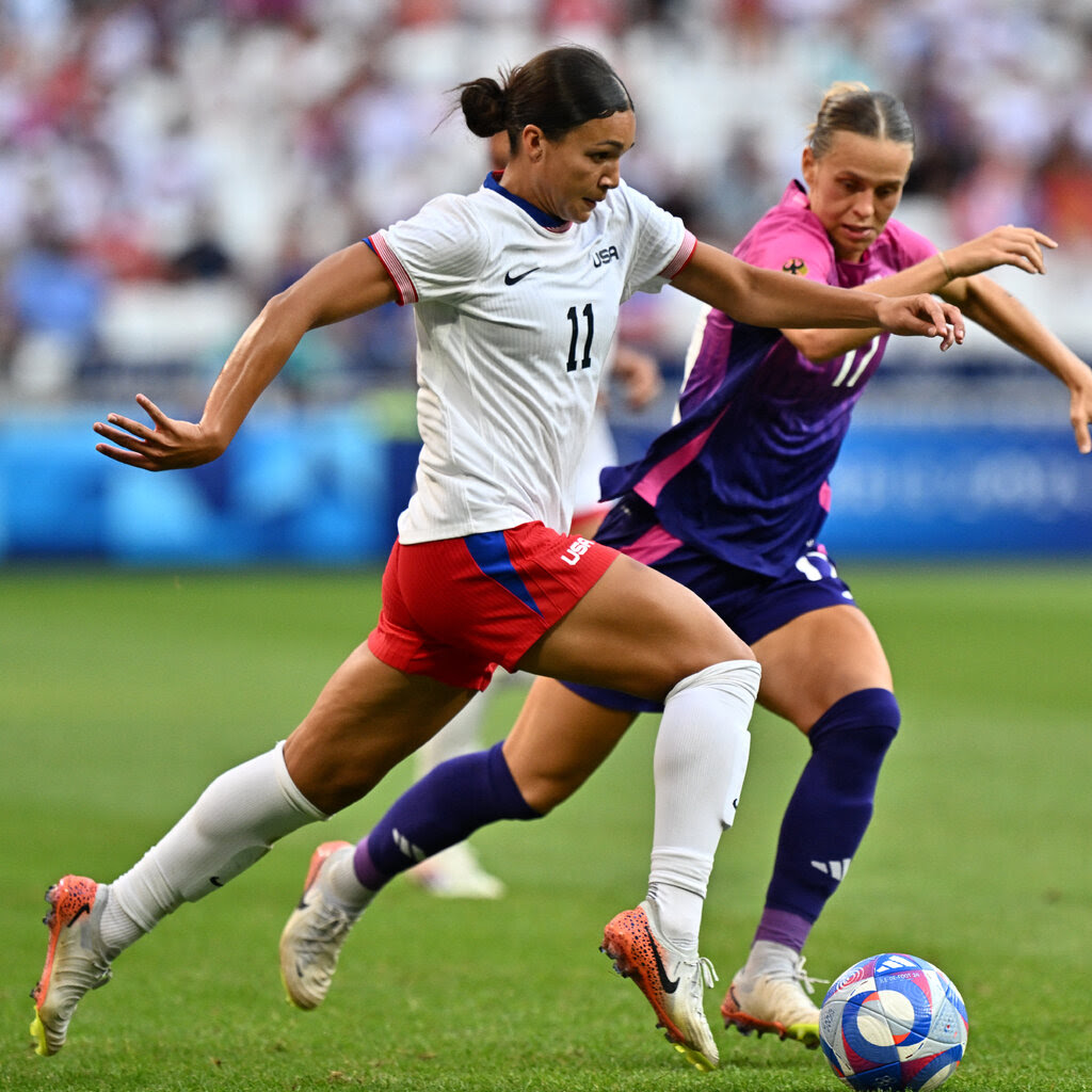 Sophia Smith wears red shorts and a white shirt. She kicks a soccer ball on the green field. Another player run behind her.