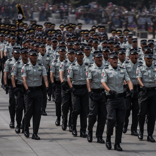 A large group marching in long lines, while wearing uniforms of black pants and gray shirts.