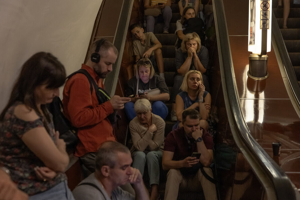 People sit and stand on an escalator. Many are checking their phones.