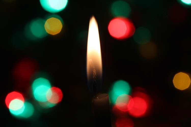 A close-up of a candle's flame against a blurred background of darkness peppered with red, turquoise, and yellow lights.