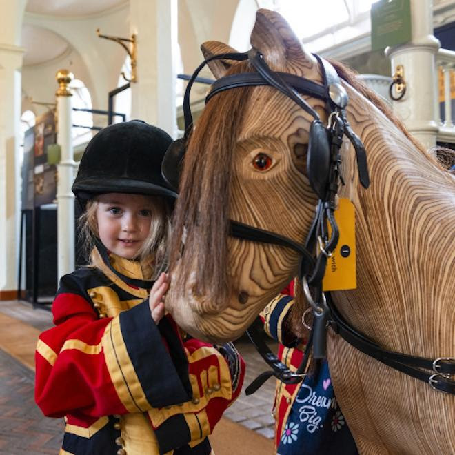 A child dressing up in livery and learning how to tack up the wooden pony at the Royal Mews