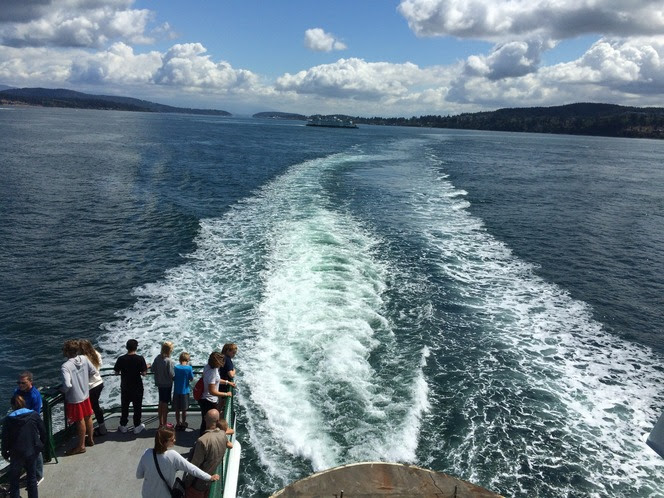 Several people on the outdoor deck on the back of a ferry in foreground with another ferry in the distance in the background