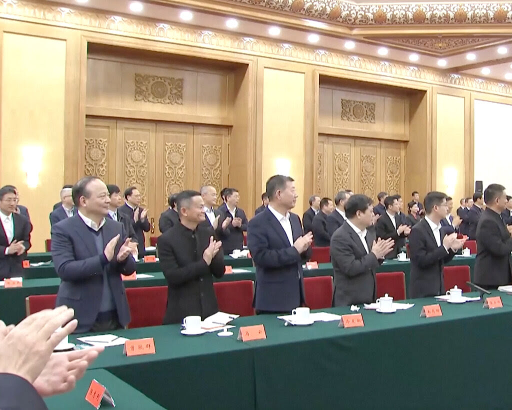 Jack Ma and other business executives standing behind a long table covered in green fabric and applauding while looking to their left. 