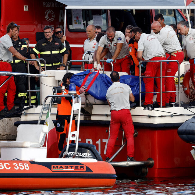 A diver in an orange jumpsuit suit and crews in gray shirts and red trousers hoist remains in a blue body bag onto a boat, as others in reflector uniforms stand nearby.