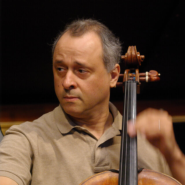 Antônio Meneses, wearing a tan shirt, plays the cello with an expression of concentration as he looks off to his right.