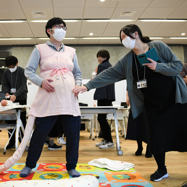 A person wears a pink smock to simulate being pregnant; a long pink cloth tube runs out of the smock and snakes on the ground. Another person stands nearby, holding a phone in one hand and placing the other hand on the simulated pregnant belly. Behind them, two people look down at baby dolls. 