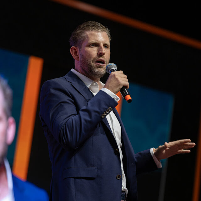 Eric Trump, con chaqueta azul y camisa blanca, sostiene un micrófono delante de una gran pantalla que proyecta su imagen.