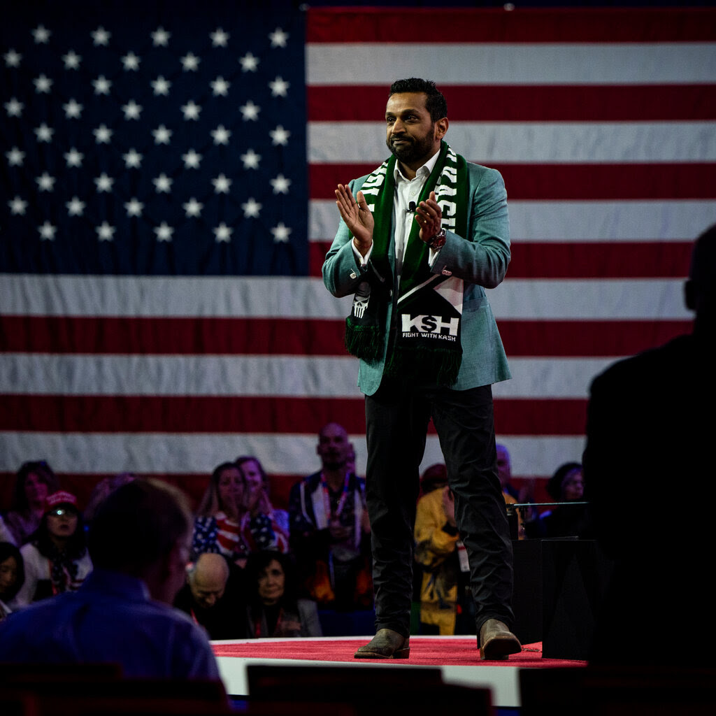 A man in a teal sports jacket and a scarf that reads “Fight With Kash” stands on a podium in front of an audience and an American flag.