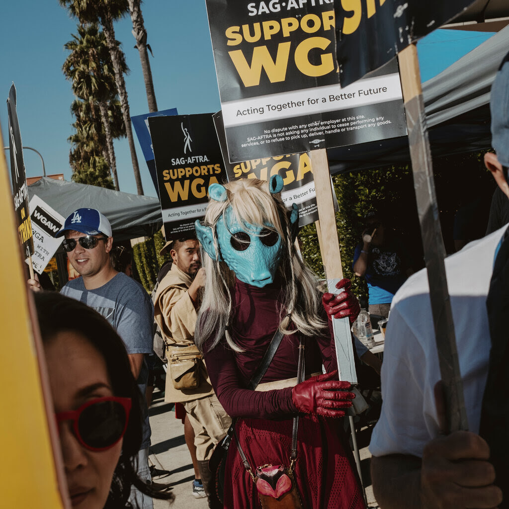 A group of people holding signs, some saying “SAG-AFTRA supports WGA.” One person is wearing a light blue mask of a creature’s face.