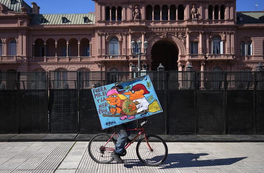 A person holding a poster rides a bicycle in front of the presidential office in Buenos Aires, Argentina. The sign says, in Spanish "Fuera Milei y la hermana."