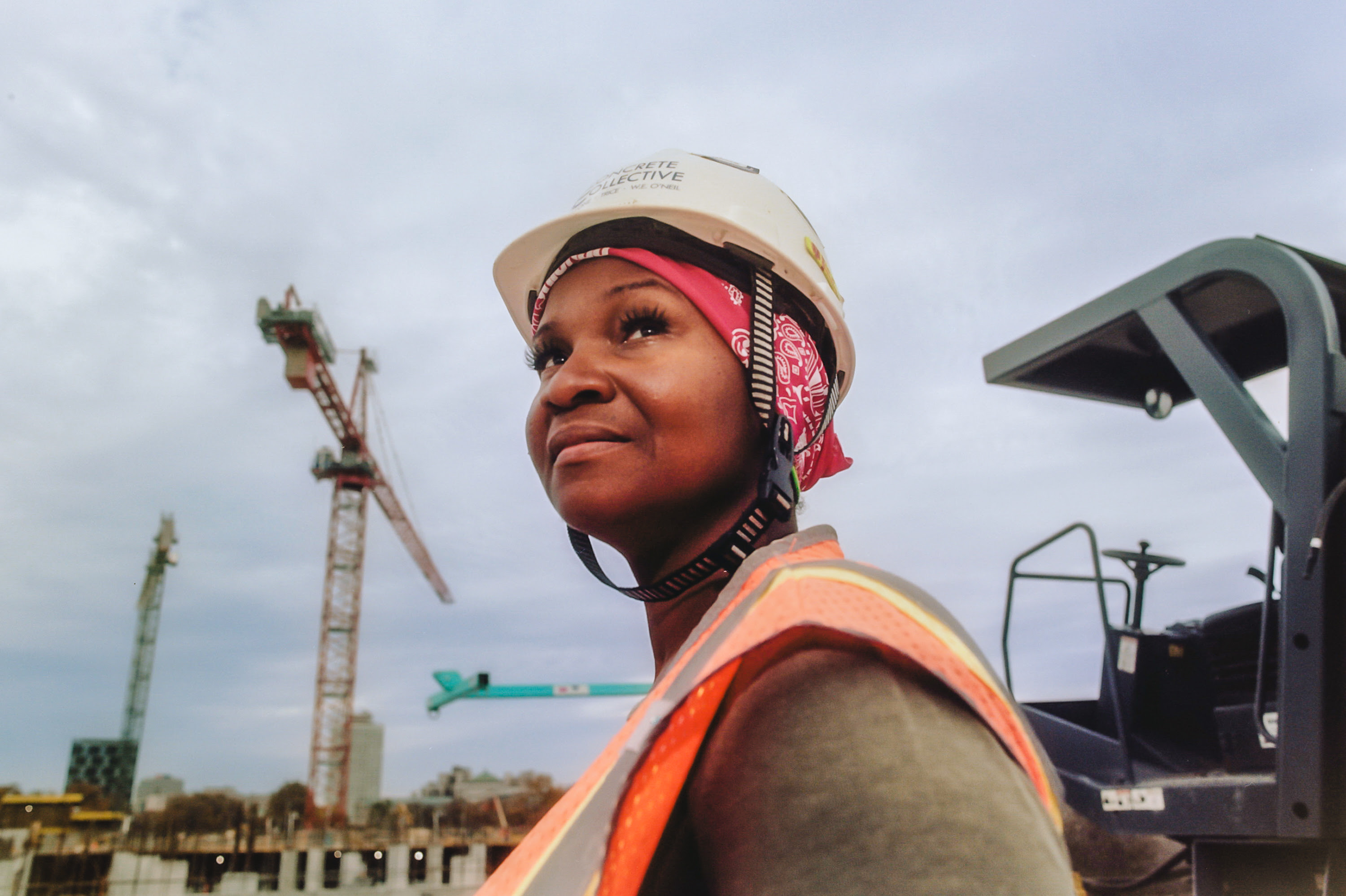 A woman with a medium skin tone is wearing a hard hat and construction vest and looking up at the sky with cranes and construction equipment in the background.