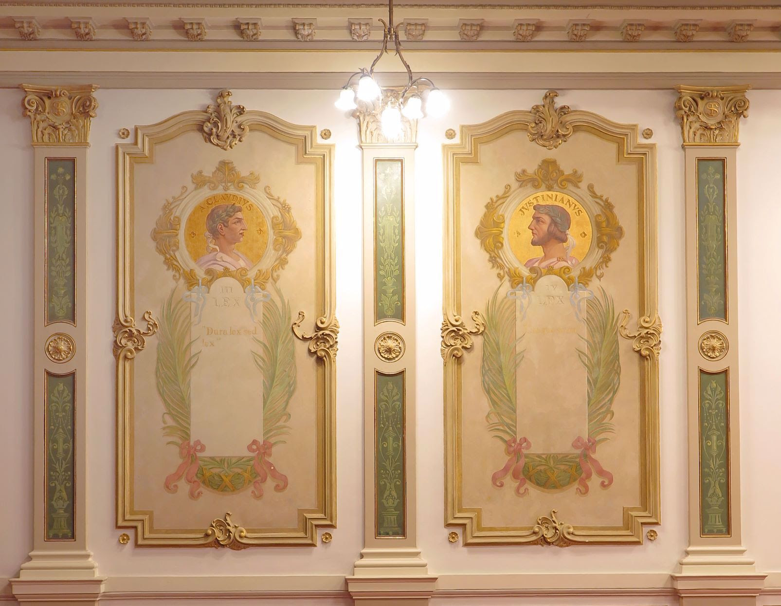 Painel decorativo em estilo clássico com dois retratos masculinos de perfil, pintados nas paredes. À esquerda, o busto de Júlio César, identificado como “CAESAR”, com a inscrição “Dura lex sed lex” abaixo. À direita, o busto de Justiniano, identificado como “IVSTINIANVS”, também acompanhado da palavra “LEX”. Ambos os retratos estão emoldurados por guirlandas, ornamentos dourados e folhas estilizadas, compondo um cenário de grande sofisticação.