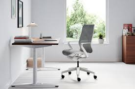 A sleek, ergonomic office chair positioned at a neat, minimalist desk setup. The chair features adjustable settings and a high backrest, surrounded by a tidy workspace with a laptop, a few pens, and a notepad, in bright natural light.