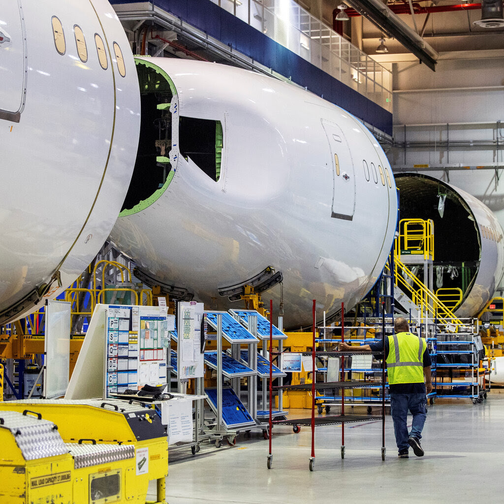 A wide frame of the inside of a Boeing 787 Dreamliner. An employee wearing a reflective construction vest is seen in the background wheeling a metal cart.