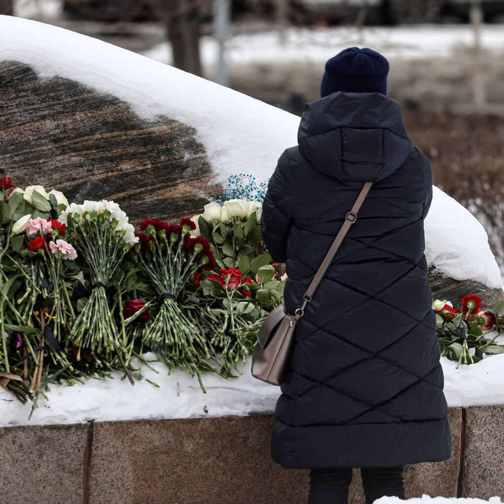 A person in a dark coat stands in front of bouquets piled at a memorial for Aleksei Navalny in Moscow.