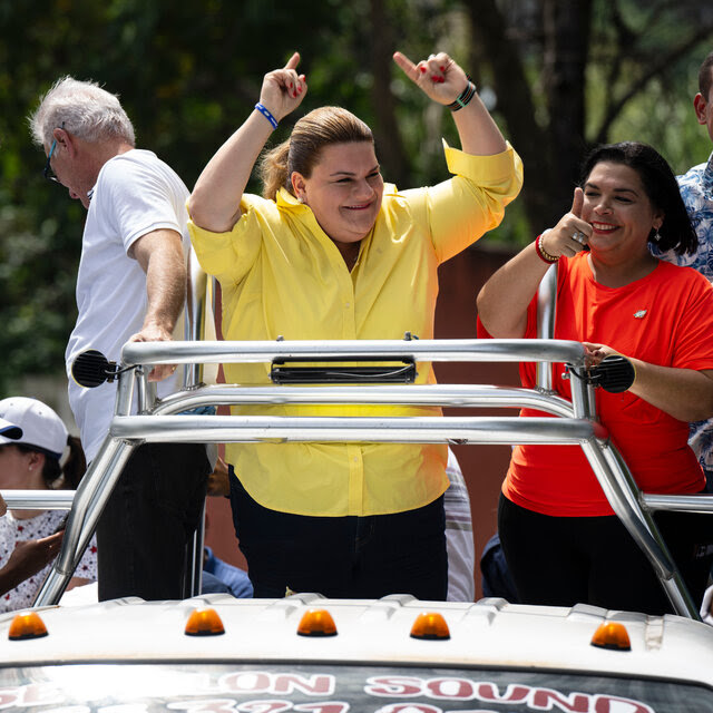  Jenniffer González-Colón, wearing a yellow shirt, smiles and stands with her hands raised and index fingers pointing upward. Nearby, a person in an orange shirt gives a thumbs-up. 