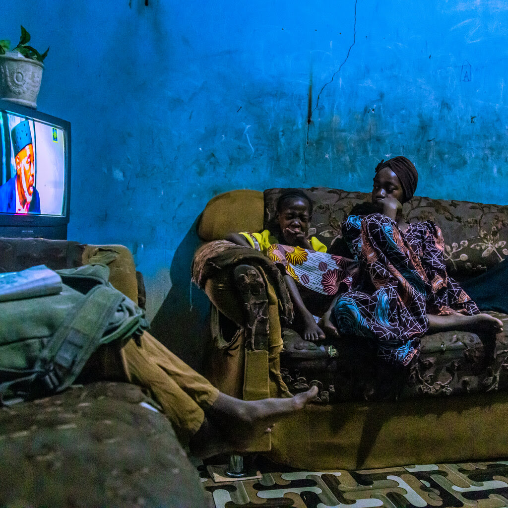 A woman, a child and a third person, only partly visible, sit on couches in a blue-walled room. A TV is on.