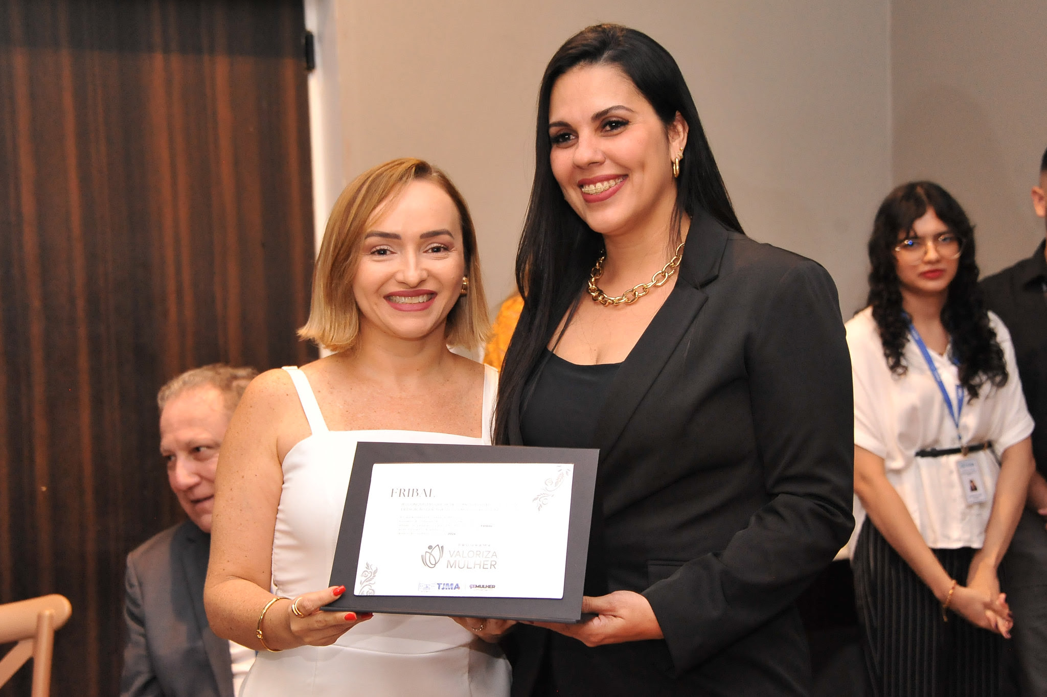 A imagem mostra duas mulheres em pé, lado a lado. A pessoa à esquerda parece estar usando uma blusa branca com um paletó por cima, e a pessoa à direita está de terno. Eles estão segurando uma placa emoldurada entre eles, de premiação do projeto Valoriza Mulher.