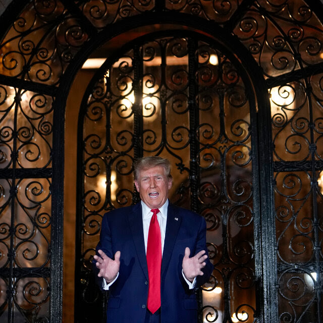 Former President Donald J. Trump in a blue suit and red tie, gesturing with his hands as he speaks in front of an ornate iron gate.