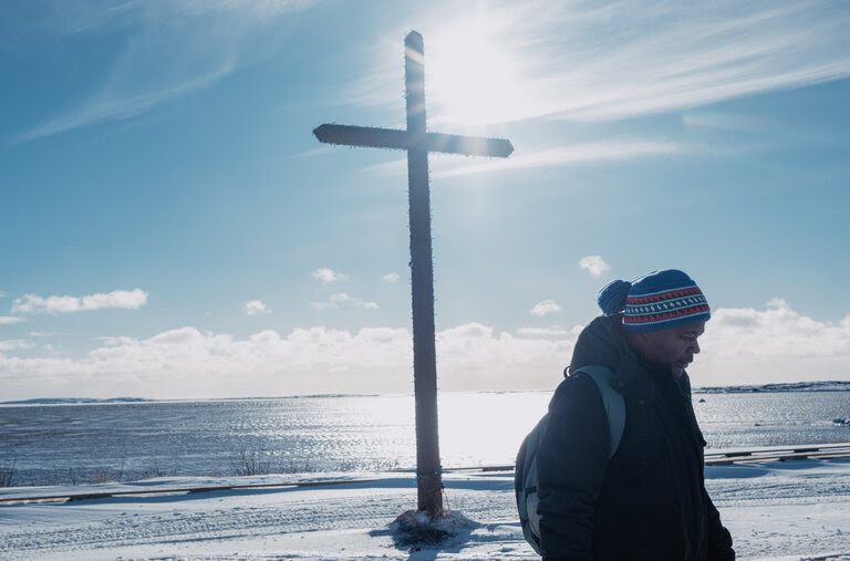 Father Gérard Tsatselam, wearing a winter coat and a wool cap, stands in front of a cross facing the Gulf of St. Lawrence.
