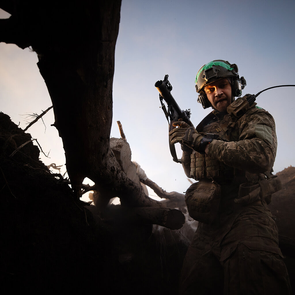 A soldier in a helmet holds a grenade launcher while standing in a trench.