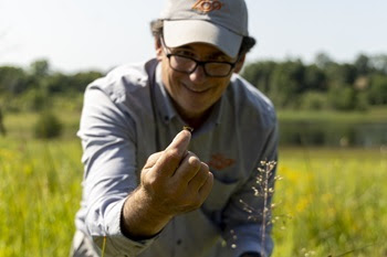 smiling man in gray shirt and ballcap and glasses kneels in a grassy field, a small orange and black butterfly resting atop his fingers