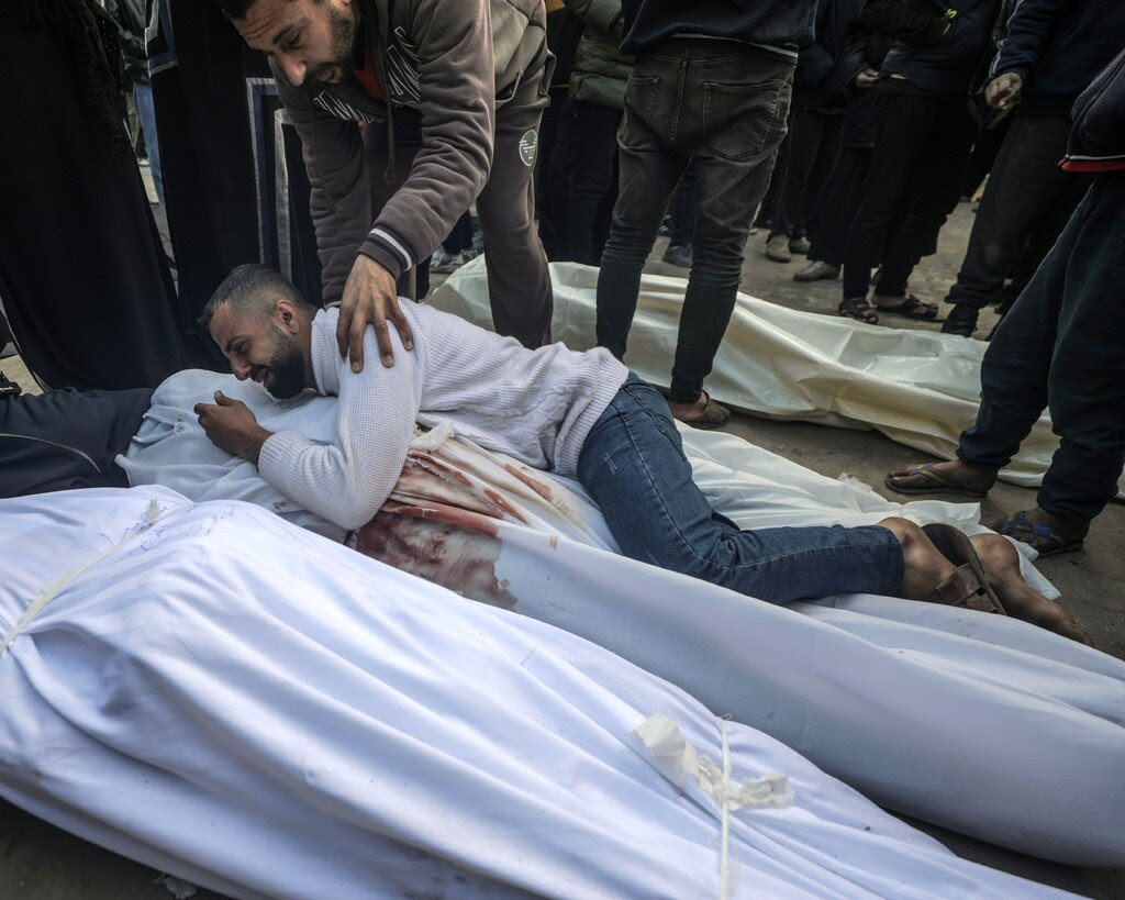 A man in mourning clutches a body wrapped in a white body bag as another man touches his shoulder. 