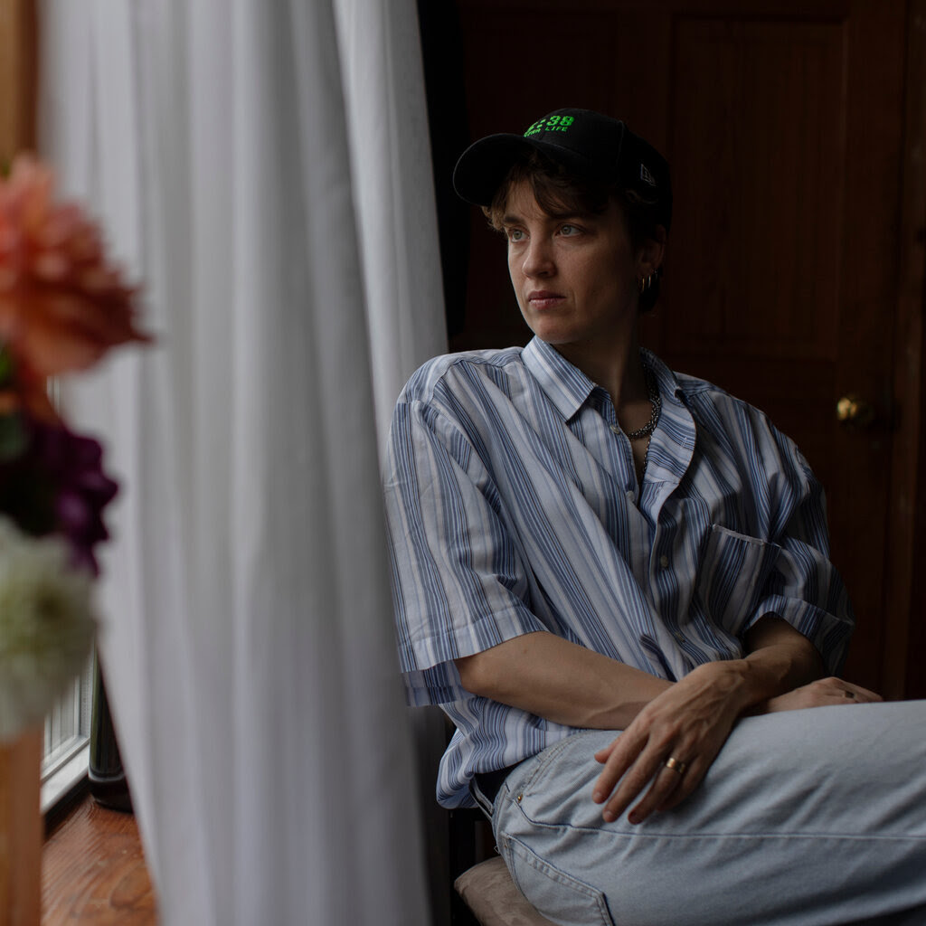 Adèle Haenel, in a blue-striped shirt and jeans and wearing a ball cap, sits and gazes out of a window. Flowers are seen in the foreground.