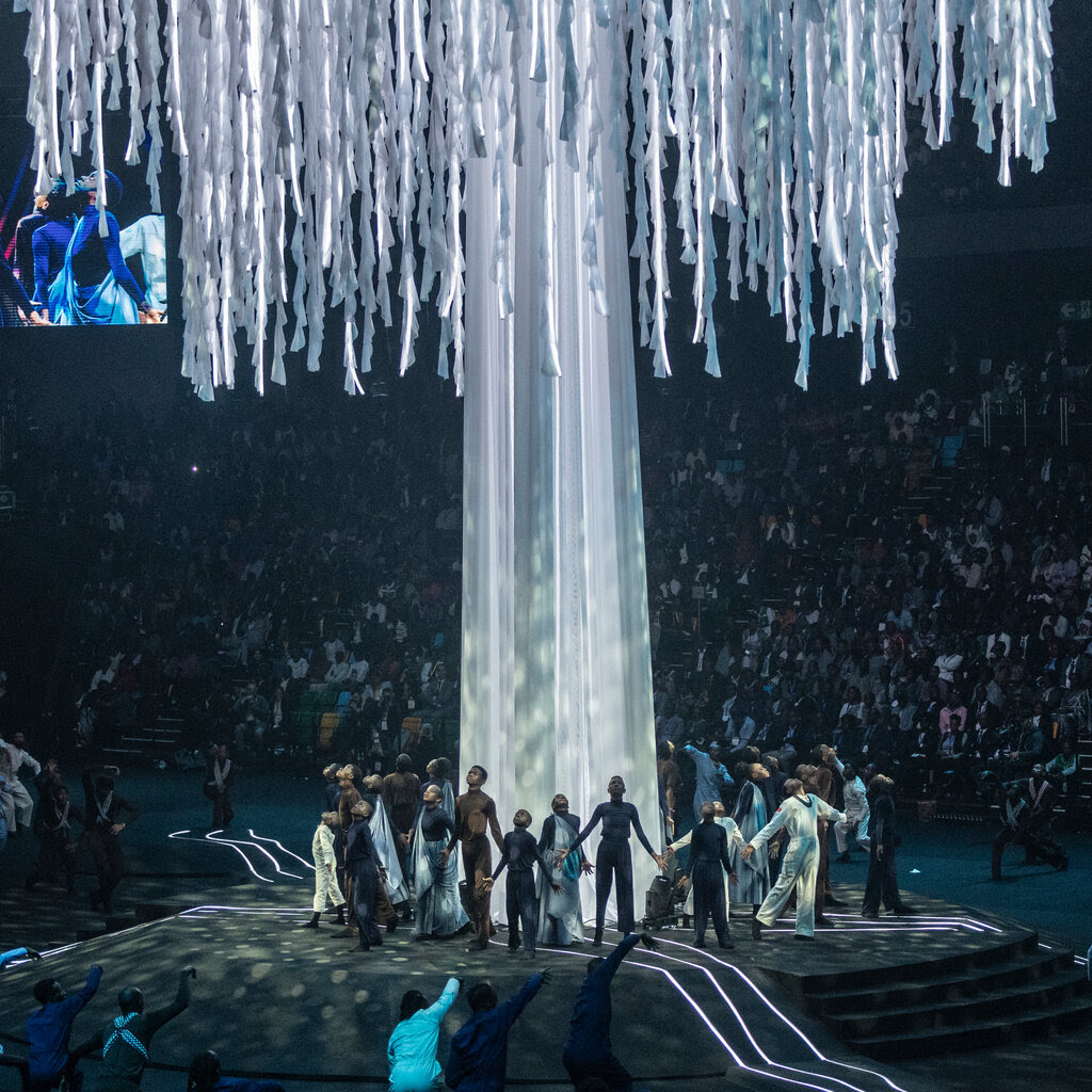 Dancers encircle a giant white “tree” on a stage in a darkened arena filled with people in the stands.