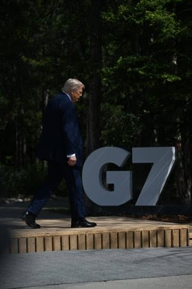 Le président Trump, vêtu d'un costume bleu, marche sur une scène en bois à l'extérieur, à côté d'une découpe blanche du G7.