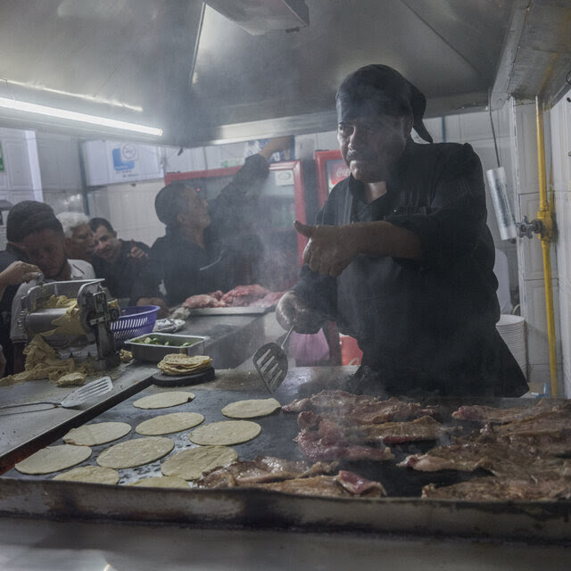 A man holds a spatula as he cooks over a hot grill with customers along the side.