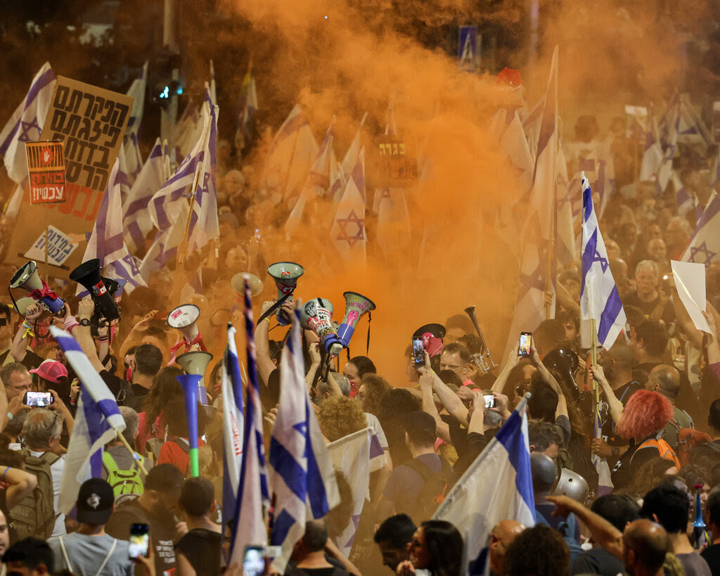 A large crowd of demonstrators holding up Israeli flags and microphones are surrounded by an orange cloud of smoke.