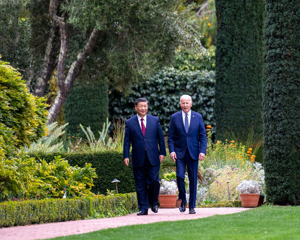 Xi Jinping and Joe Biden, both in blue suits, stroll on the lush grounds of an estate.
