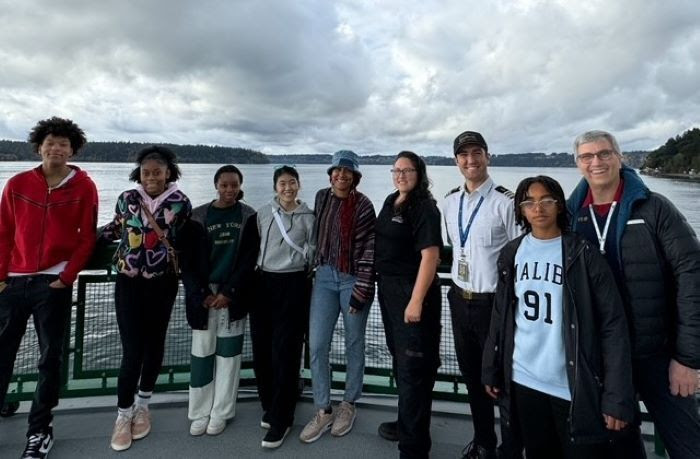 Nine people posing for a photo on the outdoor deck of a ferry with one wearing a crew uniform