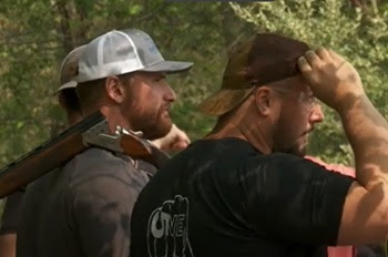 two young men with neat beards, gray-black T-shirts and baseball caps look toward the forest. One has a skeet gun open over his shoulder