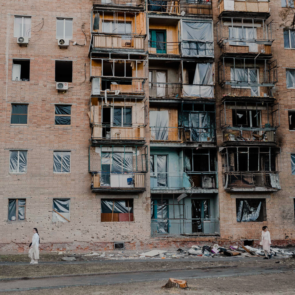 Two women wearing matching white outfits walk under a damaged brick apartment building. Debris covers the ground in their path.