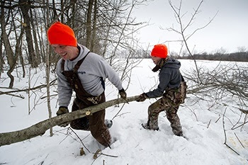 A man and a younger girl, both in bibs and winter jackets and orange knit caps, carry a long tree limb through heavy snow