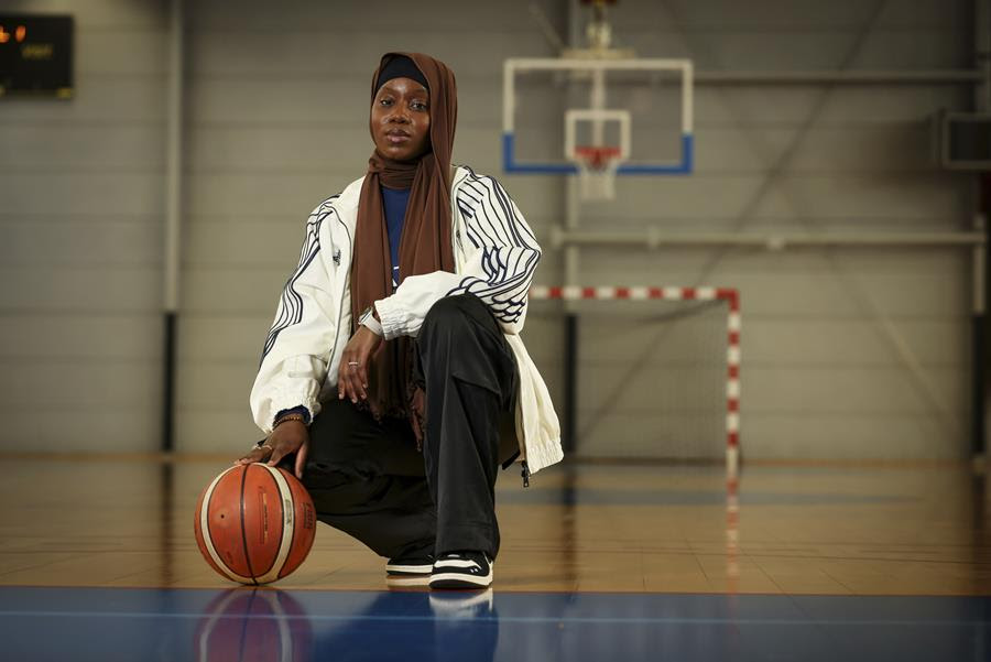 A woman wearing a headscarf and loose, athletic clothing kneels on a basketball court with her hand on a basketball.