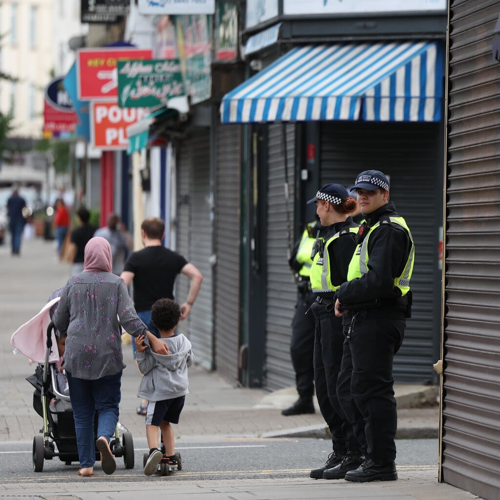 Police officers standing near shuttered shops as a woman nearby pushes a stroller with a young boy. 