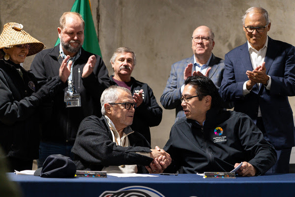 Two seated men shake hands while a group applauds behind them.