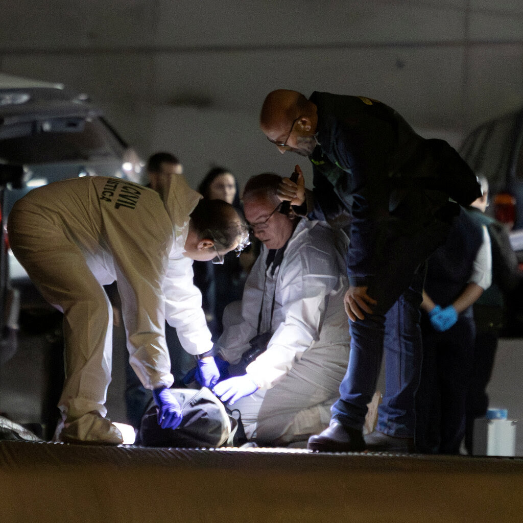 Men in blue uniforms and white hazmat suits lean over a body on the ground. They are surrounded by parked cars.