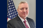 James Binniker poses in front of an American flag. Image is bordered on each side with a dark blue box