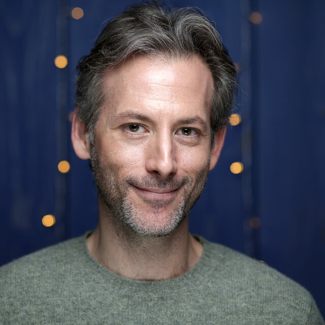 A man of graying hair and with a slight salt-and-pepper beard smiles as he poses before a blue background.