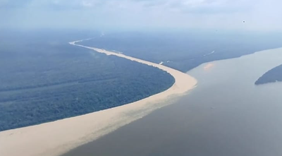 A imagem mostra uma vista aérea de dois rios com águas de cores distintas que se encontram, formando um contraste marcante entre o tom escuro e o claro, contaminado por rejeitos da mineração ilegal. Há uma densa floresta ao longo das margens e um céu parcialmente nublado no topo da cena.