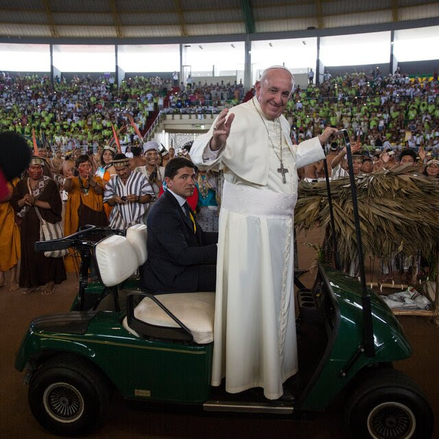 The pope in white vestments standing on a golf cart in front of a large crowd at a venue in Peru.