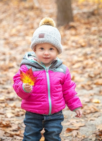 A little girl in a pink coat and pom hat holds up a red and orange maple leaf as she stands on an autumn trail