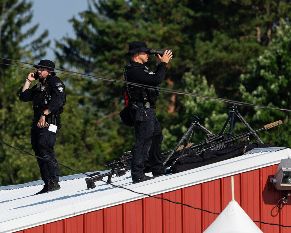 Men in black uniforms stand on a roof next to sniper rifles.